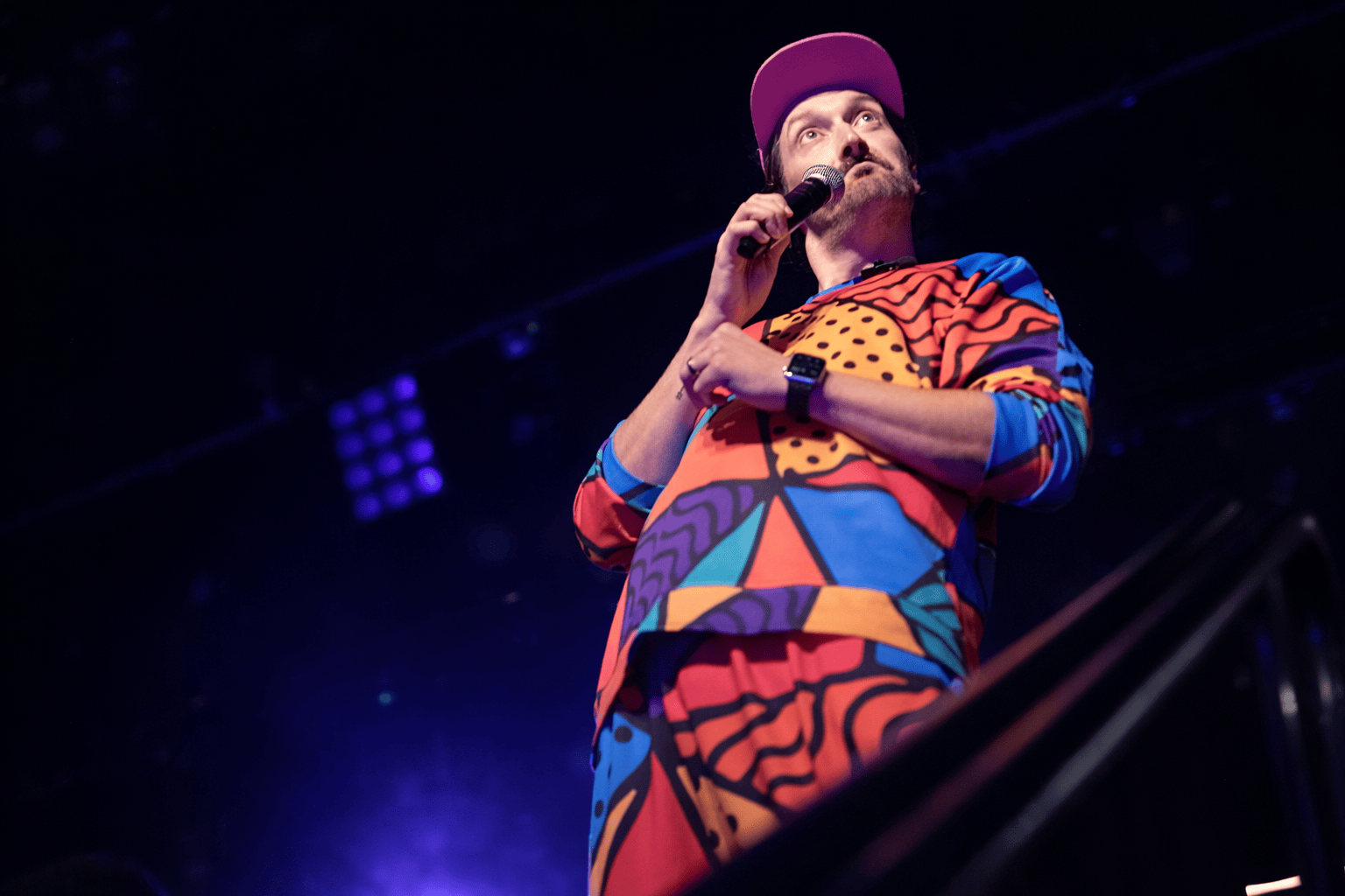 Woman in overalls and a backward cap holding a microphone with a man DJing in the background on a dark stage.